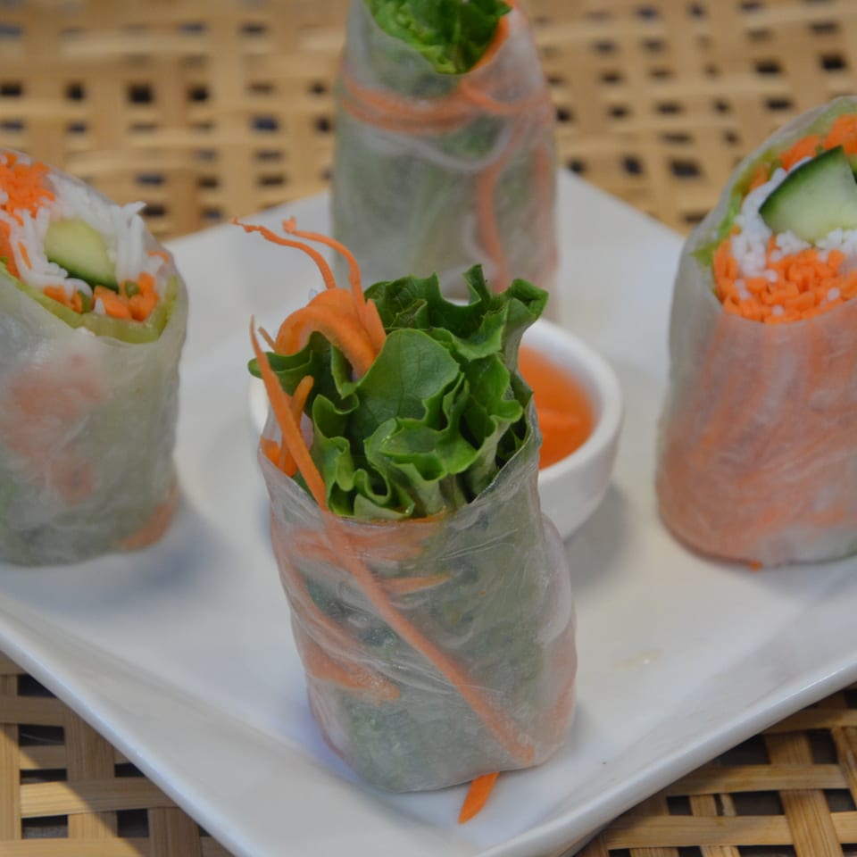 Four fresh vegetable spring rolls with lettuce, carrots, cucumber, and rice noodles arranged on a white plate with dipping sauce.