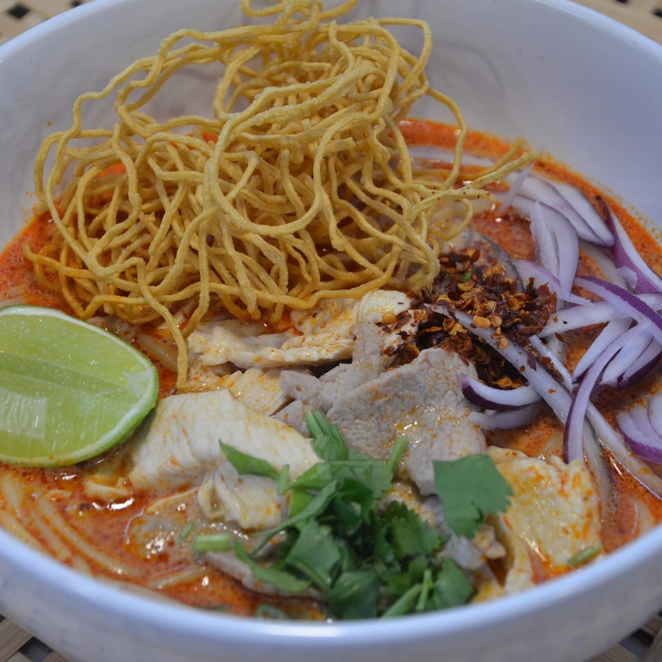 Bowl of spicy noodle soup topped with crispy noodles, sliced meat, red onions, lime, herbs, and chili flakes.