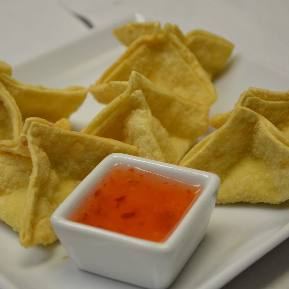 Crispy golden wontons arranged on a white plate with a square dish of bright sweet chili dipping sauce.