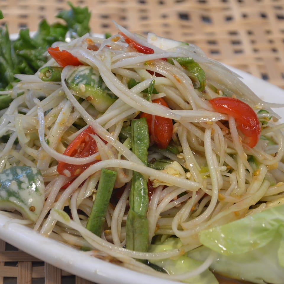 Thai-style green papaya salad with shredded papaya, tomatoes, green beans, chilies, and lettuce on a white plate.
