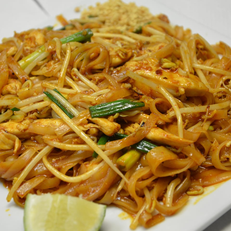 Plate of stir-fried rice noodles with chicken, bean sprouts, green onions, crushed peanuts, and lime wedge.