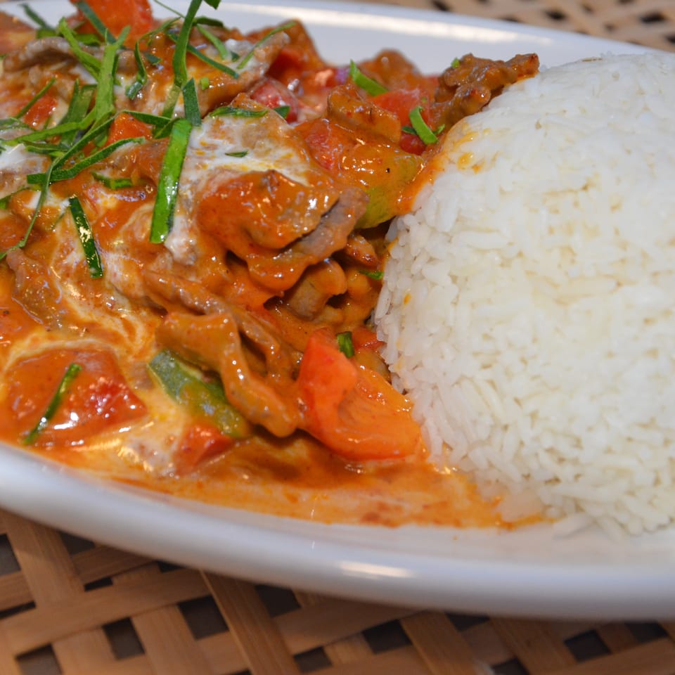 White rice served beside sliced meat and vegetables in a creamy red curry sauce, garnished with fresh herbs.