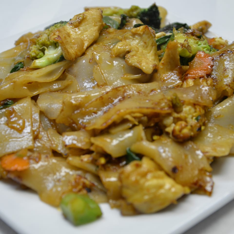 Stir-fried wide rice noodles with chicken, broccoli, carrots, and leafy greens on a white plate.