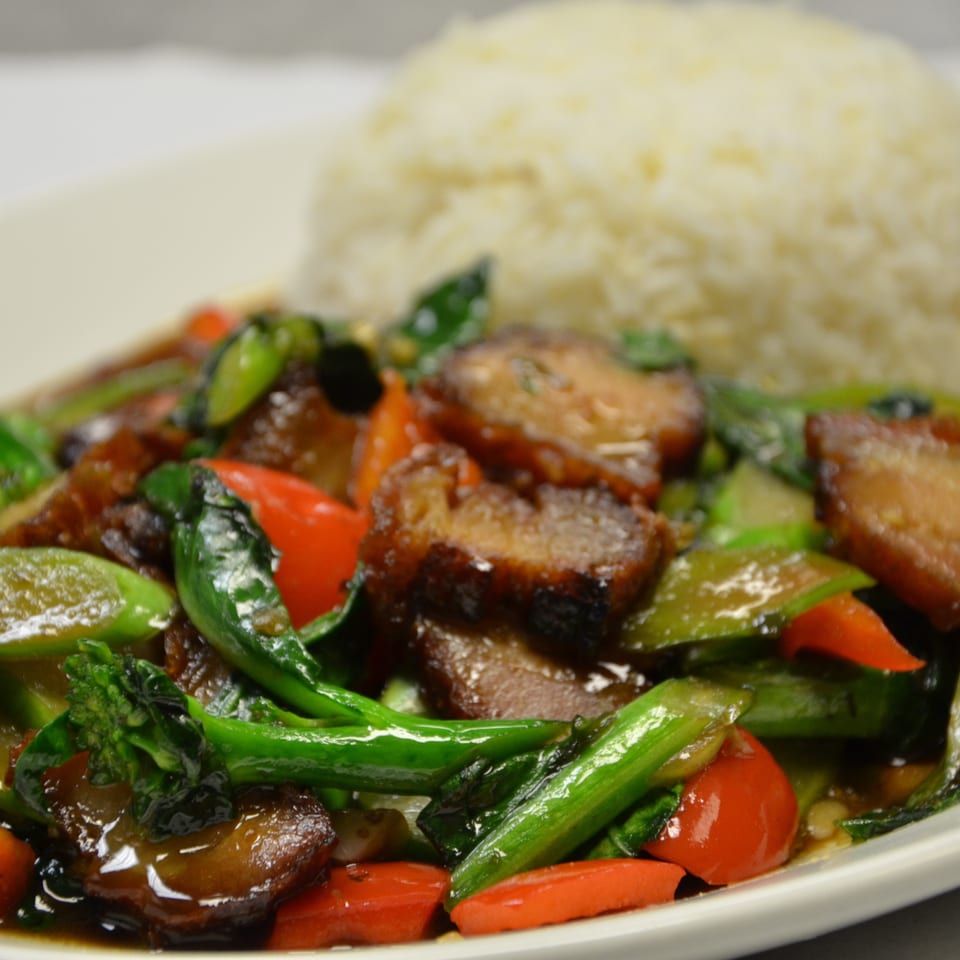 Glazed stir-fried meat and vegetables with leafy greens, peppers, and broccoli served alongside steamed rice on a white plate.