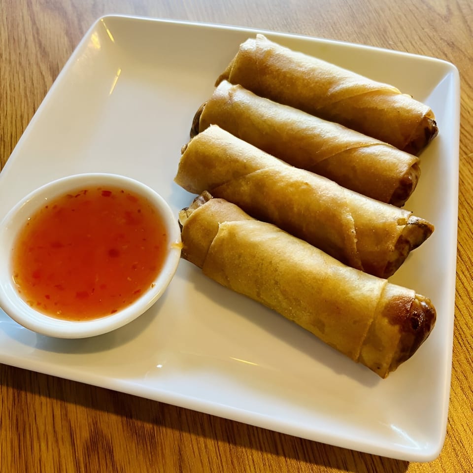 Four crispy Thai spring rolls and a small bowl of sweet chili dipping sauce on a white plate.
